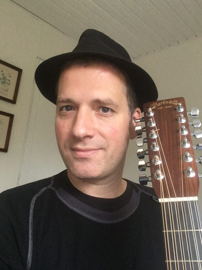 Jakob Baumgartner med sort hat, portræt med guitar bagved